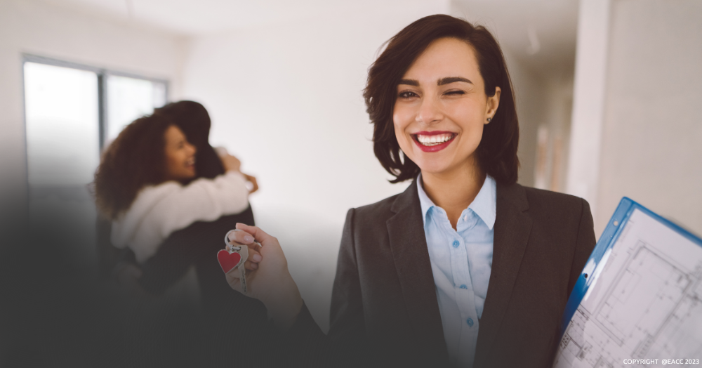 Smiling female estate agent holding keys, while behind her, a happy couple is hugging.