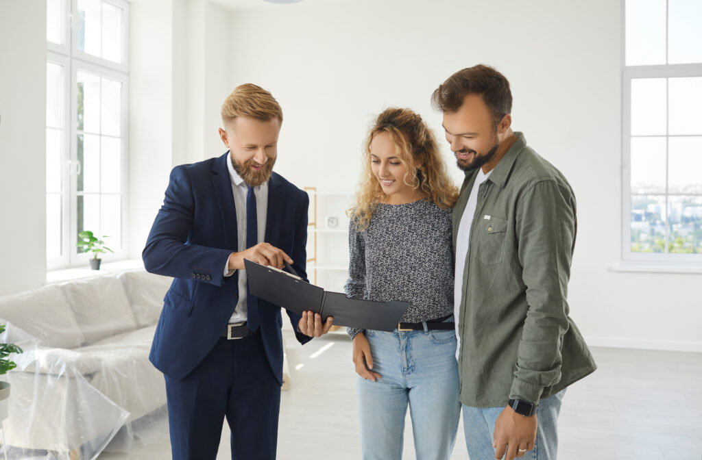 Happy married couple buying new house. Young husband and wife standing in white living room of partially furnished apartment and looking at contract that their real estate agent gives them to sign