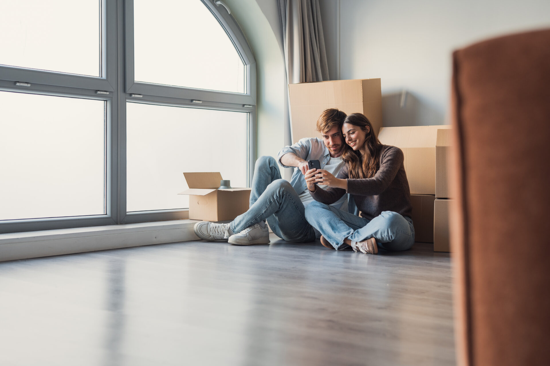 Young couple sitting on the floor in a new home surrounded by moving boxes, looking at a phone, representing first-time renters in Northwich