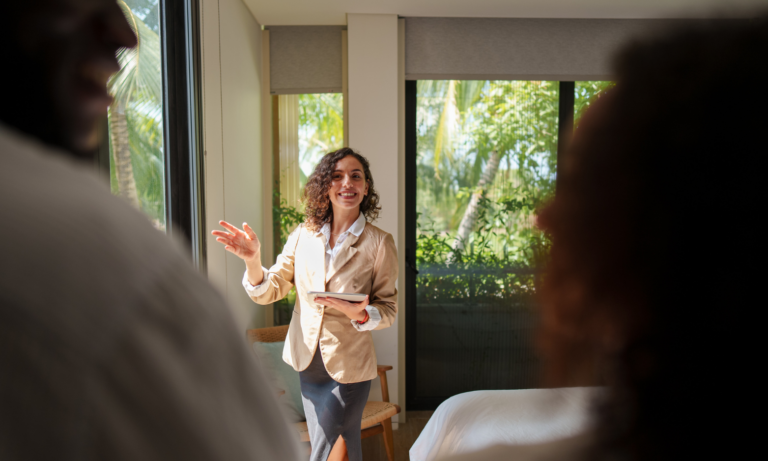 Estate agent holding a tablet and showing a bright bedroom with large windows to two prospective buyers.