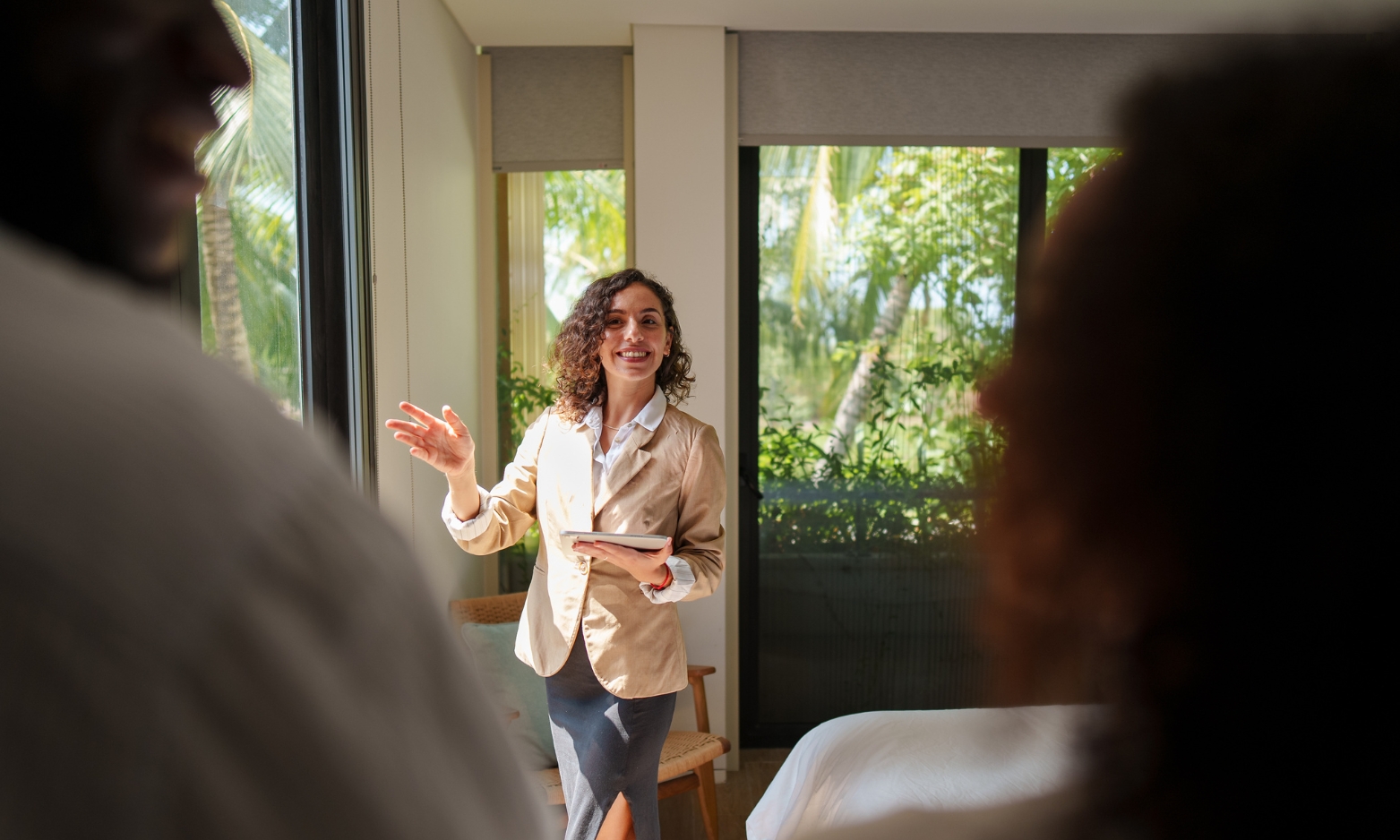 Estate agent holding a tablet and showing a bright bedroom with large windows to two prospective buyers.